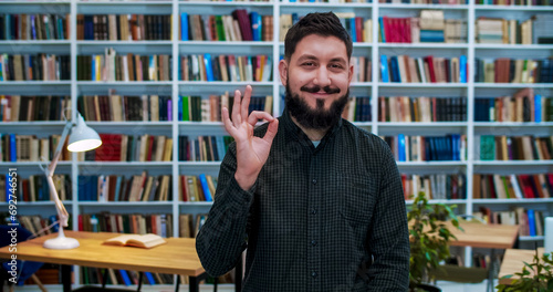 Portrait of Caucasian young man with beard smiling joyfully and showing OK gesture with hand in library room. Happy male student or teacher posing to camera and is joyful after exams. Success in study