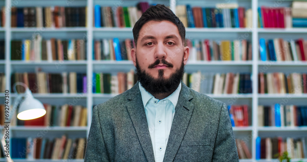 Young Caucasian businessman standing in cabinet with books shelves and looking straight to camera. Portrait of man with beard in public library or book store. Male professor. Young Caucasian businessman standing in cabinet with books shelves and looking straight to camera. Portrait of man with beard in public library or book store. Male professor.