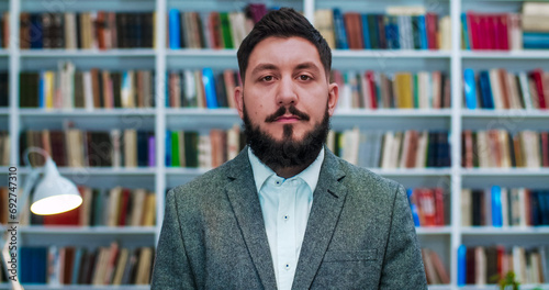 Young Caucasian businessman standing in cabinet with books shelves and looking straight to camera. Portrait of man with beard in public library or book store. Male professor. © ihorvsn