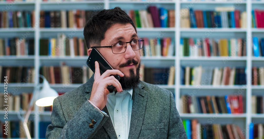 Young Caucasian businessman in glasses standing in library room and talking on mobile phone. Handsome man speaking on cellphone in bibliotheca. Telephone conversation. Male client in book store. Young Caucasian businessman in glasses standing in library room and talking on mobile phone. Handsome man speaking on cellphone in bibliotheca. Telephone conversation. Male client in book store.