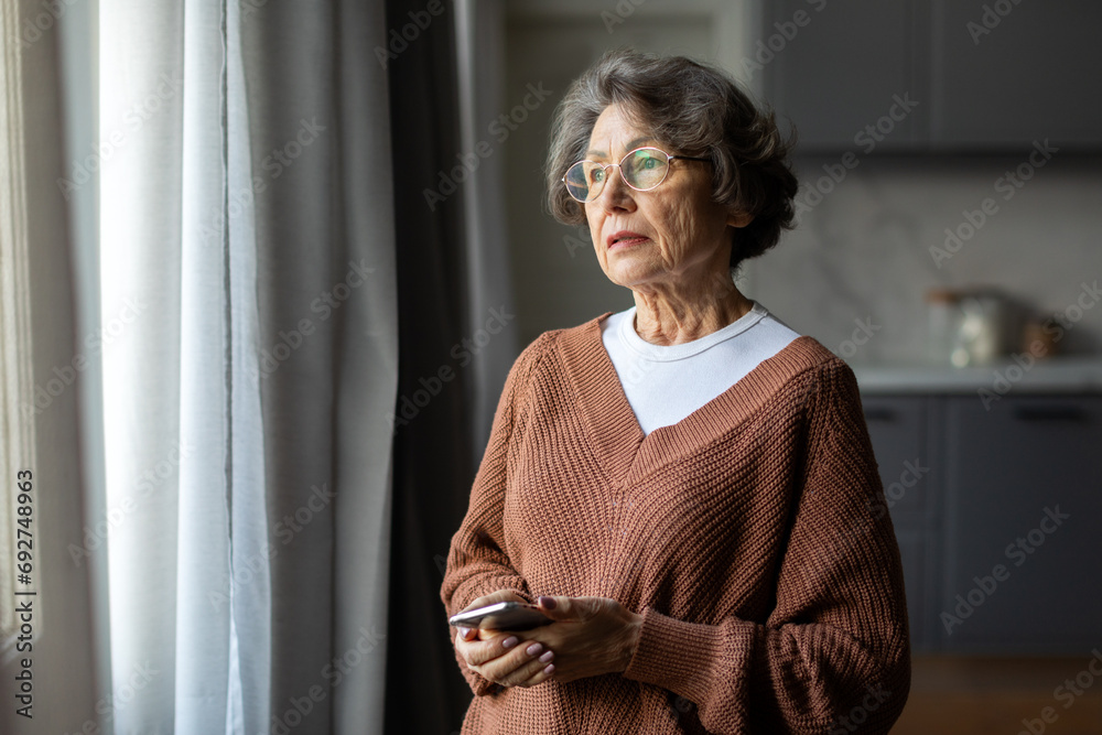 Worried depressed senior woman stand by window look away troubled, holding cellphone in hands, lady lost in sad heavy thoughts - obrazy, fototapety, plakaty 