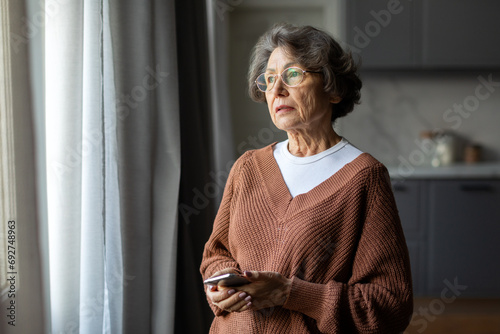 Worried depressed senior woman stand by window look away troubled, holding cellphone in hands, lady lost in sad heavy thoughts photo
