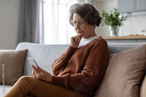 Serious senior woman using smartphone and frowning reading bad news online, sitting on couch at home, free space photo