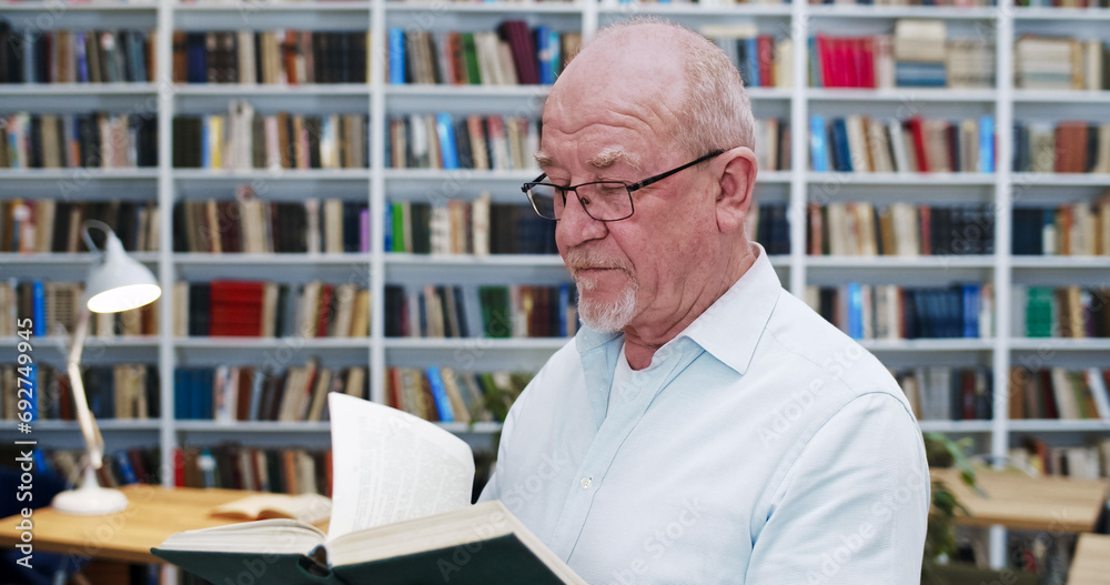 Portrait of old Caucasian bald man in glasses reading book in library and looking to camera as turning face. Close up of male professor with textbook researching in bibliotheca Scientific work concept Portrait of old Caucasian bald man in glasses reading book in library and looking to camera as turning face. Close up of male professor with textbook researching in bibliotheca Scientific work concept