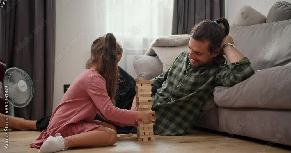 Side view of a little brunette girl in a pink dress plays with her brunette father in a checkered green shirt the board game and when she pulls out one of the bricks the Jenga tower falls