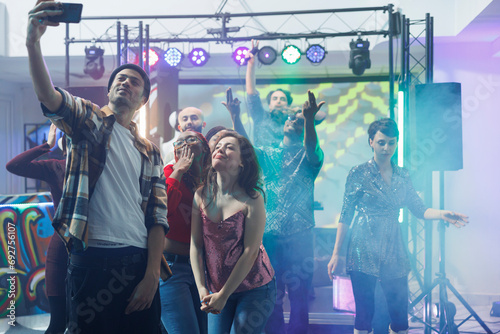 Man clubber taking selfie with women while partying at nightclub discotheque. Young friends posing for mobile phone photo while dancing and having fun on club dancefloor illuminated with lights