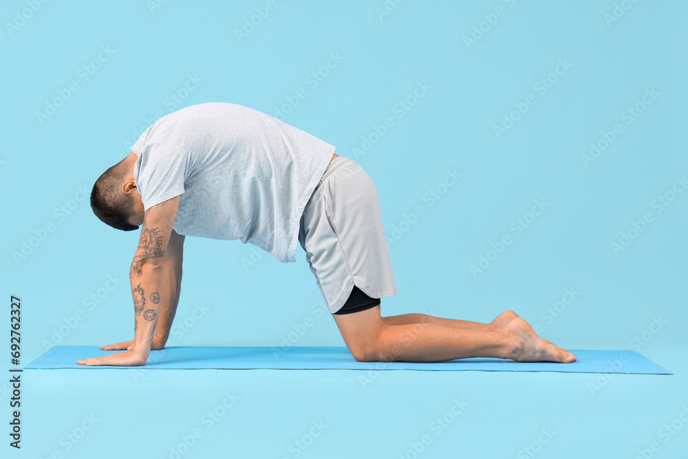 Young man doing yoga on mat against blue background - obrazy, fototapety, plakaty 
