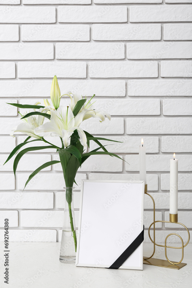 Blank funeral frame, burning candles and vase with lily flowers on table near light brick wall Blank funeral frame, burning candles and vase with lily flowers on table near light brick wall