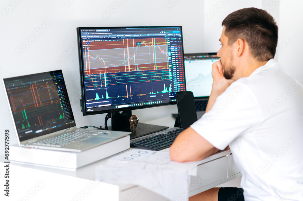 Thoughtful caucasian male crypto trader sitting at home at his work desk in front of monitors with graphs, thinking about cryptocurrency strategy, income, losses, analyzing Thoughtful caucasian male crypto trader sitting at home at his work desk in front of monitors with graphs, thinking about cryptocurrency strategy, income, losses, analyzing