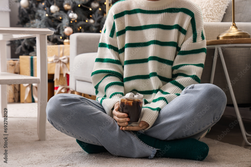 Obraz premium Woman holding glass of hot chocolate with marshmallows in festive living room, closeup