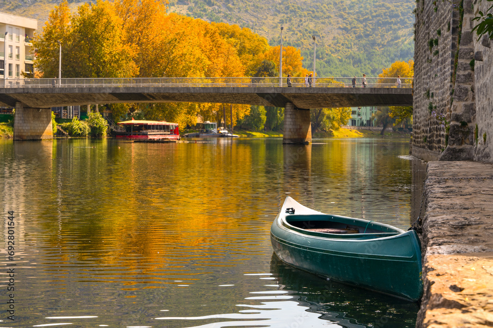 Obraz premium Golden autumn landscape in historical tourist city with bridge Trebinje Bosnia and Herzegovina snow-free cityscape, boat near ancient stone fortress, reflected in surface of river, yellow trees Balkan
