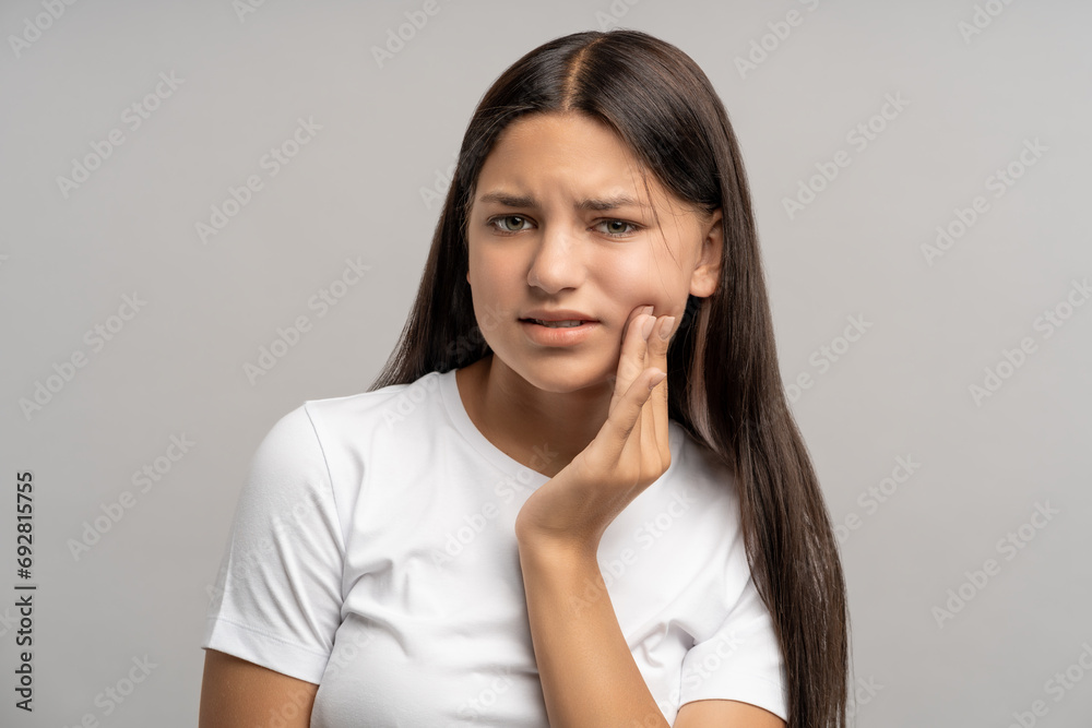 Unhappy teenage girl touching cheek with hand with painful face expression caused by toothache, dental pain, pulpitis, caries, periodontal inflammation. Teenager in need of stomatological treatment Unhappy teenage girl touching cheek with hand with painful face expression caused by toothache, dental pain, pulpitis, caries, periodontal inflammation. Teenager in need of stomatological treatment