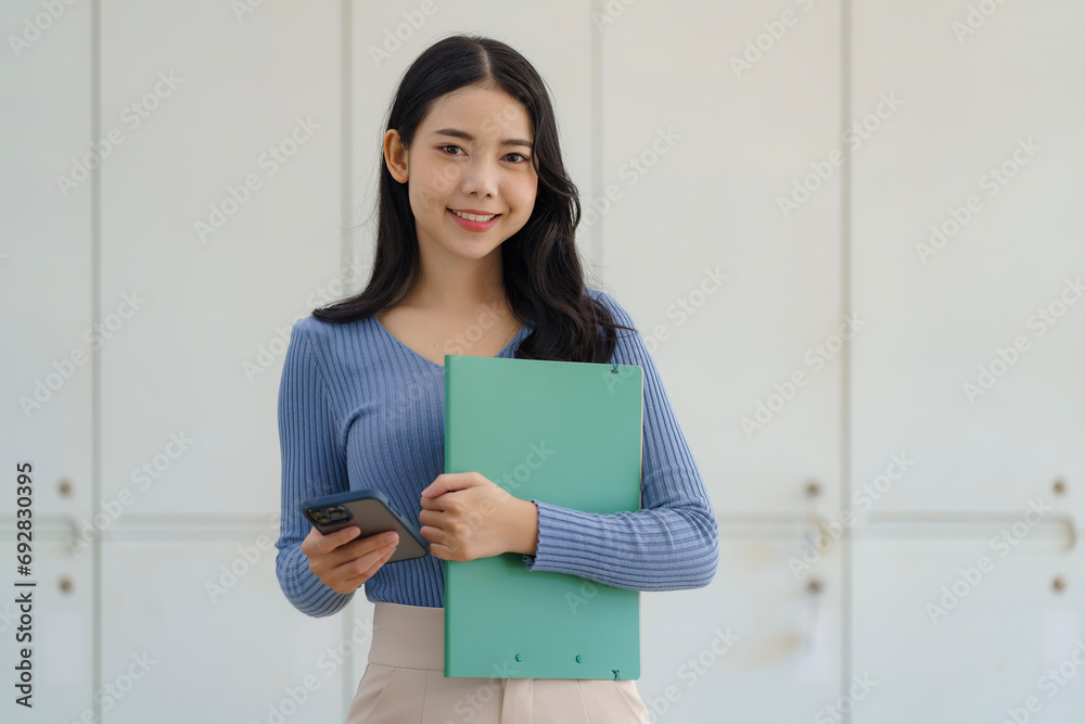 Young beautiful Asian business woman holding a folder and a smart phone while standing at the workplace. Young beautiful Asian business woman holding a folder and a smart phone while standing at the workplace.
