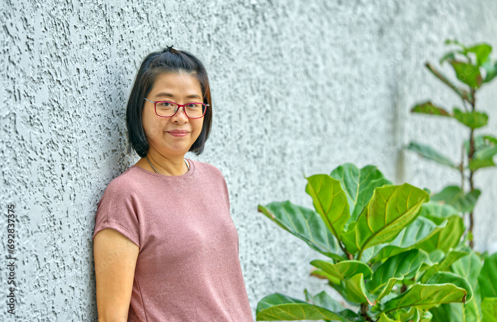 Portrait Asian middle-aged woman standing against the wall in the garden, wearing eyeglasses, looking at the camera, natural light, smiling, black short hair, healthy middle-aged woman. Portrait Asian middle-aged woman standing against the wall in the garden, wearing eyeglasses, looking at the camera, natural light, smiling, black short hair, healthy middle-aged woman.