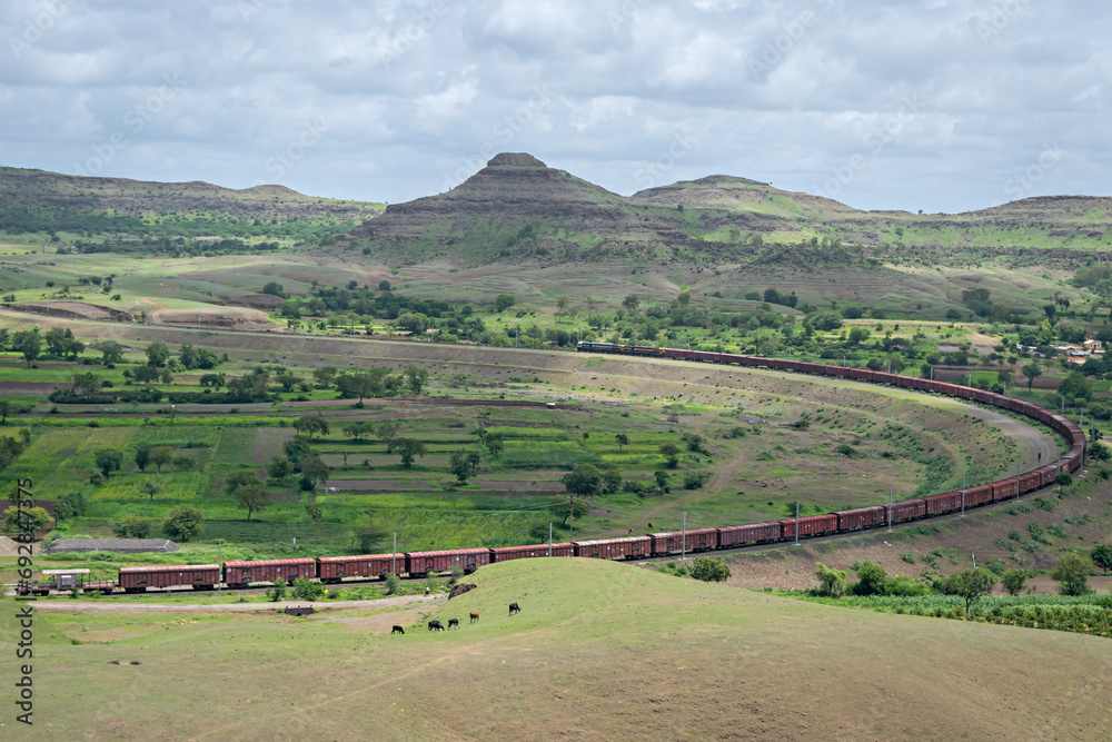Long goods freight train passing through a big curve in hilly railway line with monsoon clouds hovering over , at Daundaj, Maharashtra, India.