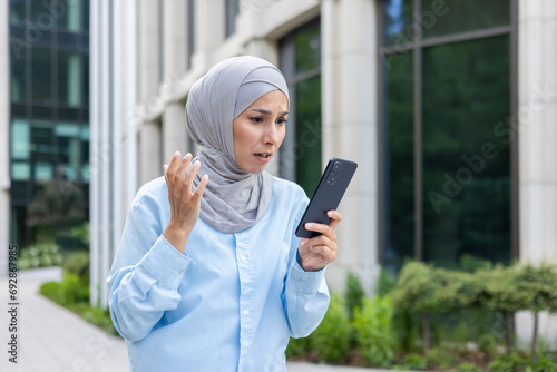 Worried arab woman in hijab scared by bad news on phone, standing on street near office, shocked by what she read.