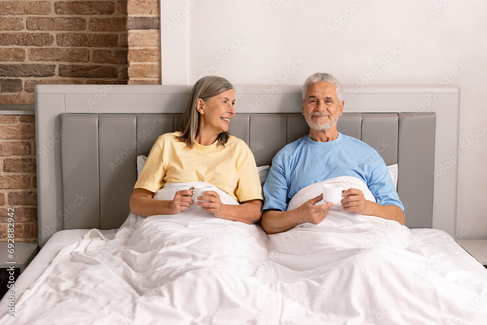 Fototapeta premium Happy senior couple sitting, drinking coffee and talking together in their bed.