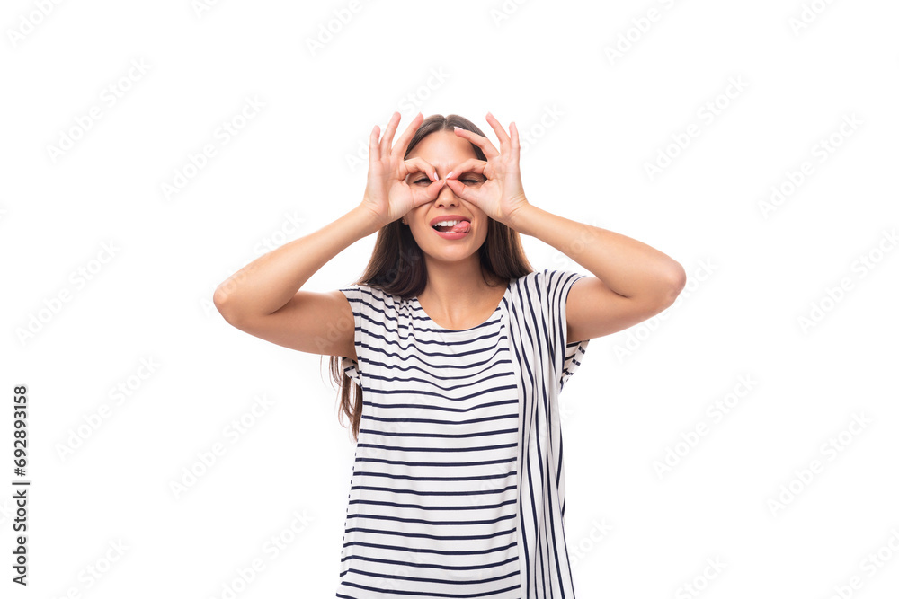 young cheerful brunette lady with straight hair in glasses is dressed in a striped t-shirt and jeans makes a grimace on a white background with copy space young cheerful brunette lady with straight hair in glasses is dressed in a striped t-shirt and jeans makes a grimace on a white background with copy space
