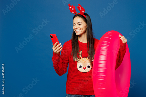 Traveler Latin woman in red Christmas sweater hold rubber ring mobile cell phone isolated on plain blue background. Tourist travel abroad in free time getaway. Air flight trip Happy New Year concept. #692924339