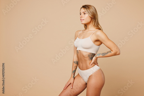 Side view young calm nice lady woman with slim body perfect skin wear nude top bra lingerie stand akimbo hold hand on waist isolated on plain pastel light beige background. Lifestyle diet fit concept. © ViDi Studio