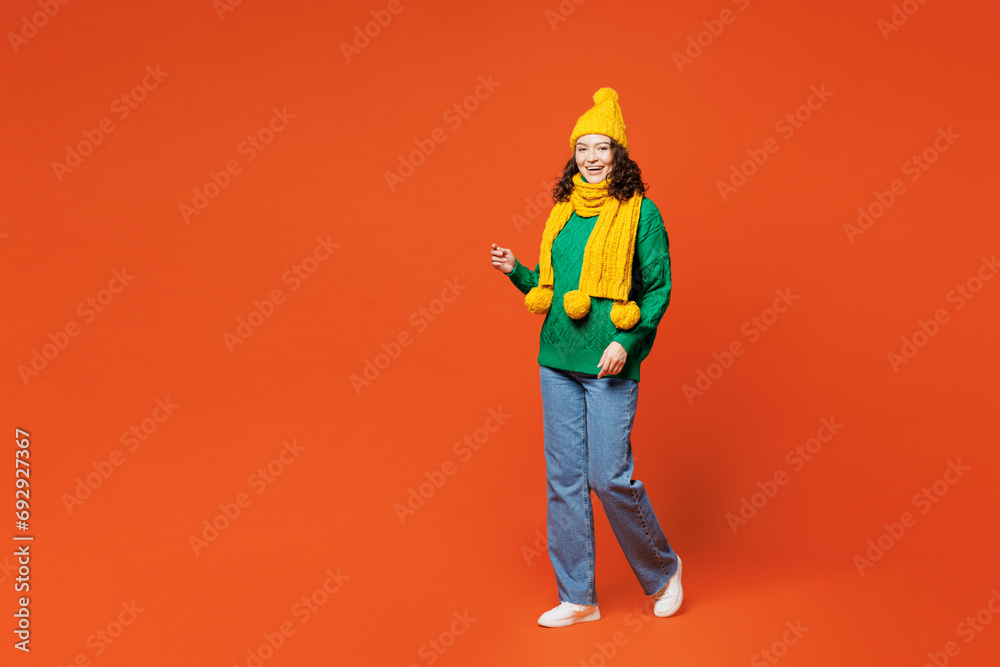 Full body side view young smiling cheerful happy woman she wear green knitted sweater yellow hat scarf walk go look camera isolated on plain orange red background studio portrait. Lifestyle concept. Full body side view young smiling cheerful happy woman she wear green knitted sweater yellow hat scarf walk go look camera isolated on plain orange red background studio portrait. Lifestyle concept.