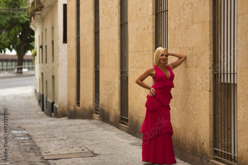 Mature woman, beautiful, blonde, wearing a pink flamenco suit with ruffles and polka dots, happy and smiling, posing leaning against a wall on a lonely street. Concept beauty, flamenco, maturity. © Manuel
