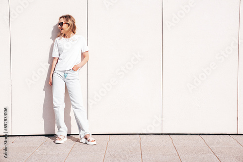 Portrait of beautiful smiling model in sunglasses. Female dressed in summer hipster white T-shirt and jeans. Posing near wall in the street. Funny and positive woman having fun outdoors