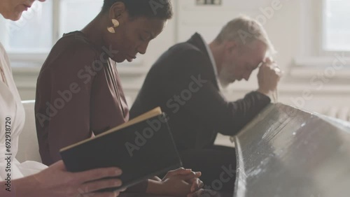 Lens flared slowmo shot of group of young and adult male and female multiracial parishioners looking down while praying, sitting on bench during sermon in church