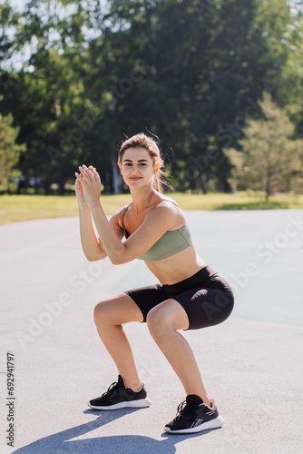 Woman smiling during squat exercise in sunny park setting. Cheerful girl at outdoor training. Sport, fitness...