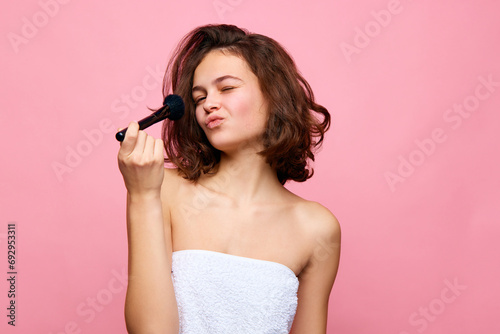 Pretty young brunette girl with short curly hair, doing makeup, applying blush, face powder against pink background. Concept of natural female beauty, skin care, cosmetology and cosmetics