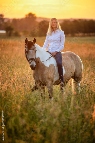 Young woman sits on her horse bareback in a meadow with the sunrise behind her.