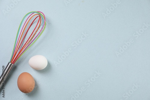 Colorful whisk and raw eggs on light blue background, flat lay. Space for text
