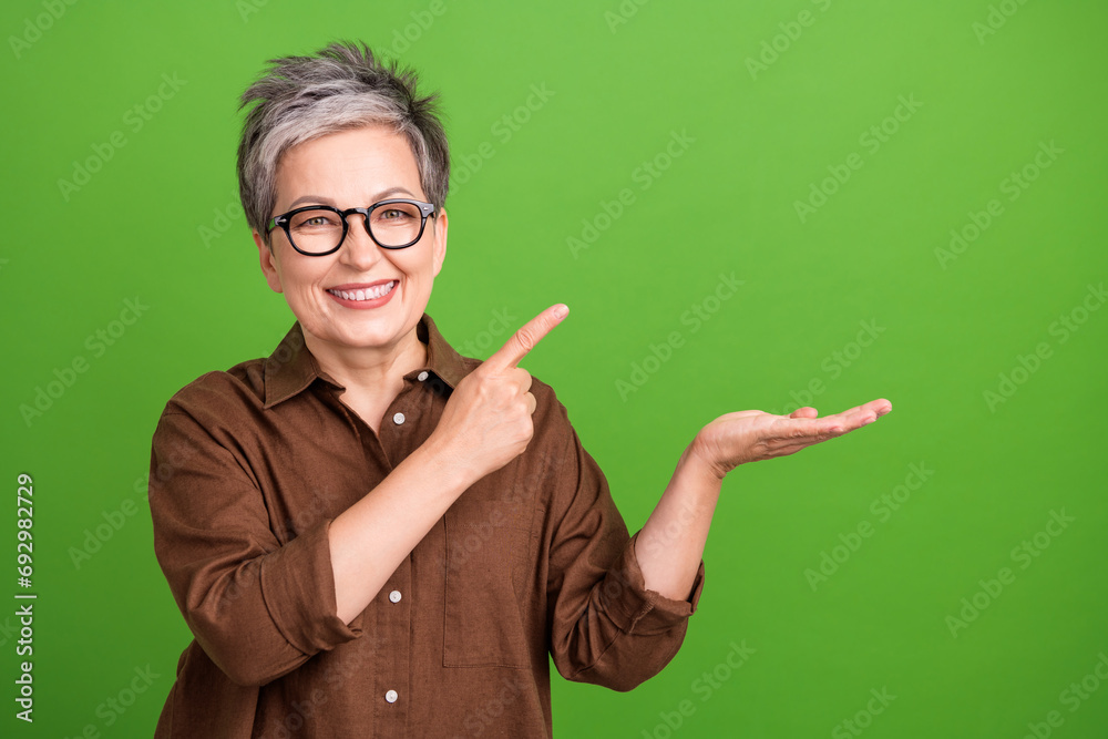 Photo of cheerful woman with short hairstyle wear brown shirt directing at offer on arm empty space isolated on green color background Photo of cheerful woman with short hairstyle wear brown shirt directing at offer on arm empty space isolated on green color background