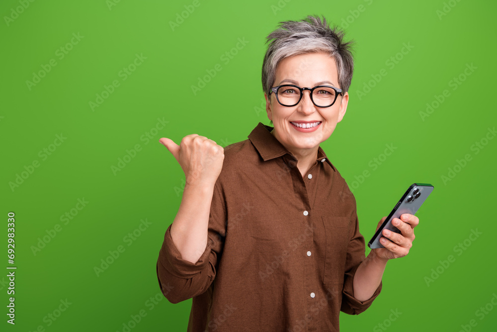 Portrait of nice person dressed brown clothes in eyewear hold smartphone indicating at sale empty space isolated on green color background Portrait of nice person dressed brown clothes in eyewear hold smartphone indicating at sale empty space isolated on green color background
