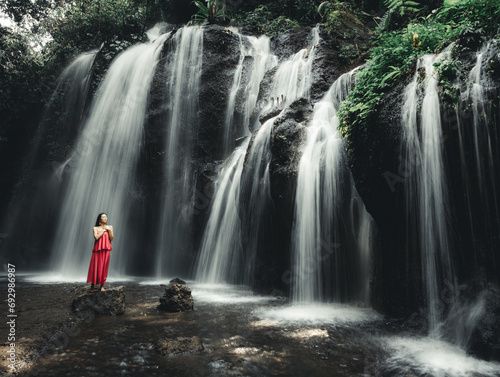 Asian woman posing near waterfall. Nature and environment. Motion water. Travel lifestyle. Young woman wearing long red dress. Slim body. Copy space. Yeh Bulan waterfall  Bali