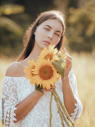 Caucasian woman walking in a field and holding a bouquet of sunflowers. Portrait of charming woman wearing white dress. Summer time. Lifestyle concept.