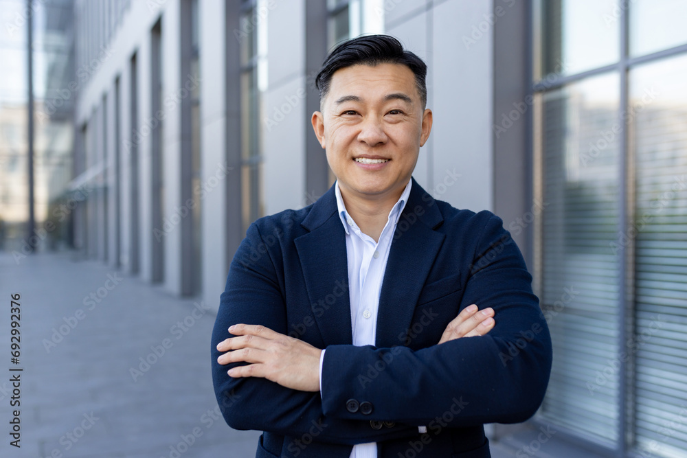 Close-up portrait of a smiling young businessman standing in a suit outside an office center, crossing his arms over his chest and looking confidently into the camera Close-up portrait of a smiling young businessman standing in a suit outside an office center, crossing his arms over his chest and looking confidently into the camera