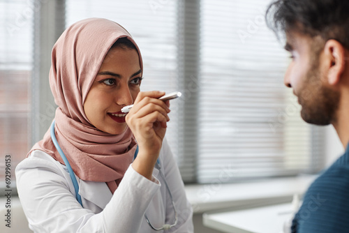 Medium close up of woman medical practitioner wearing hijab checking eyes of male patient with medical penlight photo