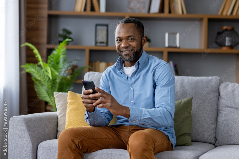 Portrait of adult mature man, African American man smiling and looking at camera with phone in hand, happy sitting on couch in living room at home, using app on smartphone.