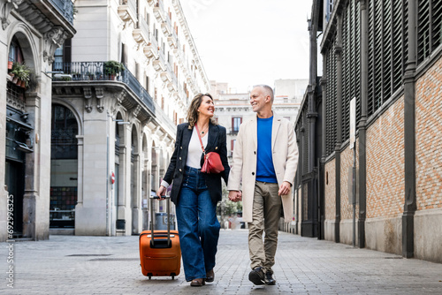 Affectionate mid adult tourist couple walking together with luggage in a european city. Lovely happy senior man and woman traveling with a suitcase visiting streets of old town.