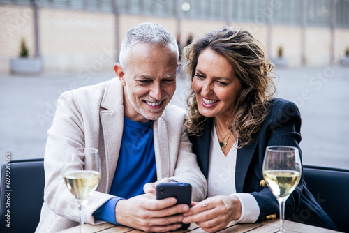 Joyful mature man and woman sharing smartphone while drinking wine in city at outdoors terrace bar. Cheerful handsome mid age couple using mobile phone together at a table restaurant.