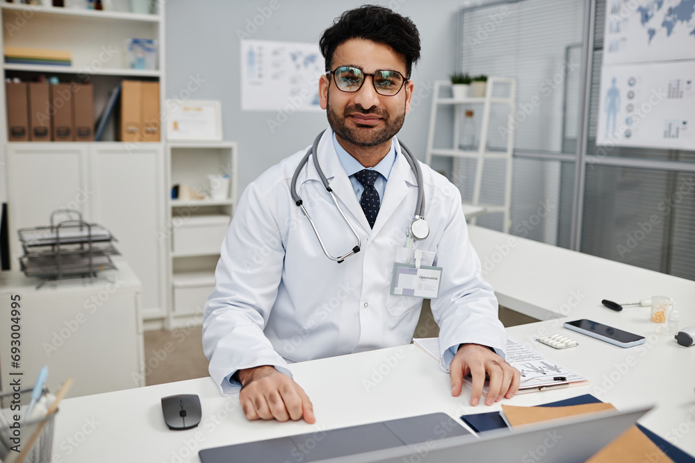 Portrait of Middle Eastern male doctor in glasses looking at camera while sitting at workplace in modern clinic office Portrait of Middle Eastern male doctor in glasses looking at camera while sitting at workplace in modern clinic office