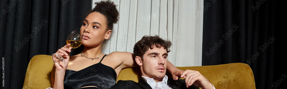 Obraz premium elegant african american woman drinking wine and sitting with man in suit on velvet sofa, banner