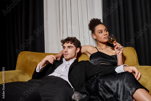 elegant young african american woman drinking wine and sitting with man in suit on velvet sofa © LIGHTFIELD STUDIOS