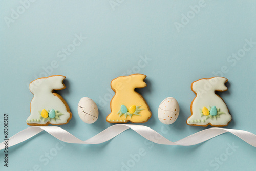 Easter homemade baked gingerbread or cookies with pattern of icing, bunny and eggs. White chocolates candies - Easter eggs. Decor with white ribbon. Blue background, top view, copy space