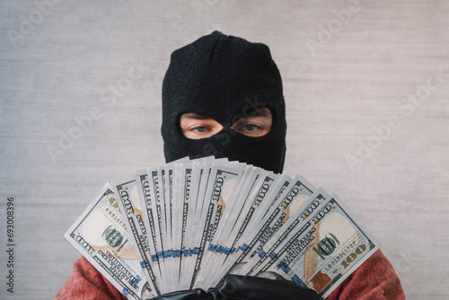 the concept of crime and money theft. Male thief with balaclava on his head holding a handful of dollar bills in front of his eyes