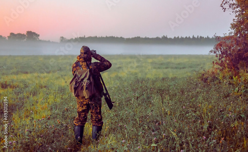  Man with gun stands on the edge of an autumn field in search of hunting object at dawn.