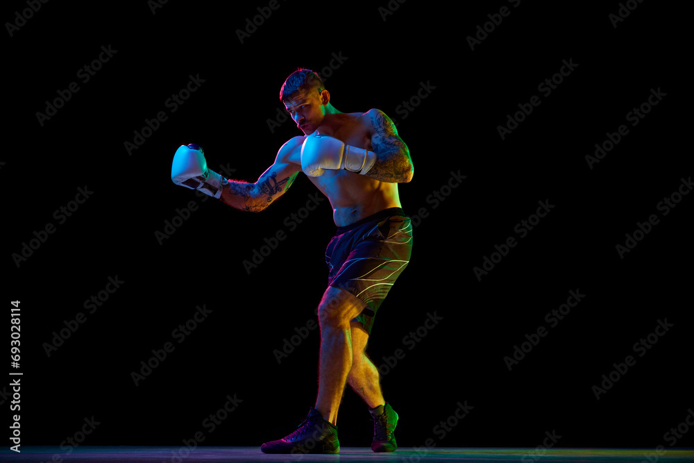 Full-length image of young athletic man, muscular, shirtless boxer in motion, fighting, practicing hooks isolated over black background in neon. Concept of sport, combat sport, martial arts, strength