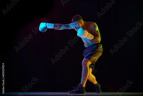 Obraz na płótnie Strong, muscular young man, boxing athlete in gloves, training, fighting isolated over black background in neon light