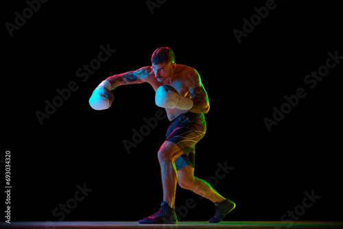 Full-length of shirtless young man with muscular strong body, boxer in motion, training isolated on black background in neon light. Concept of professional sport, combat sport, martial arts, strength